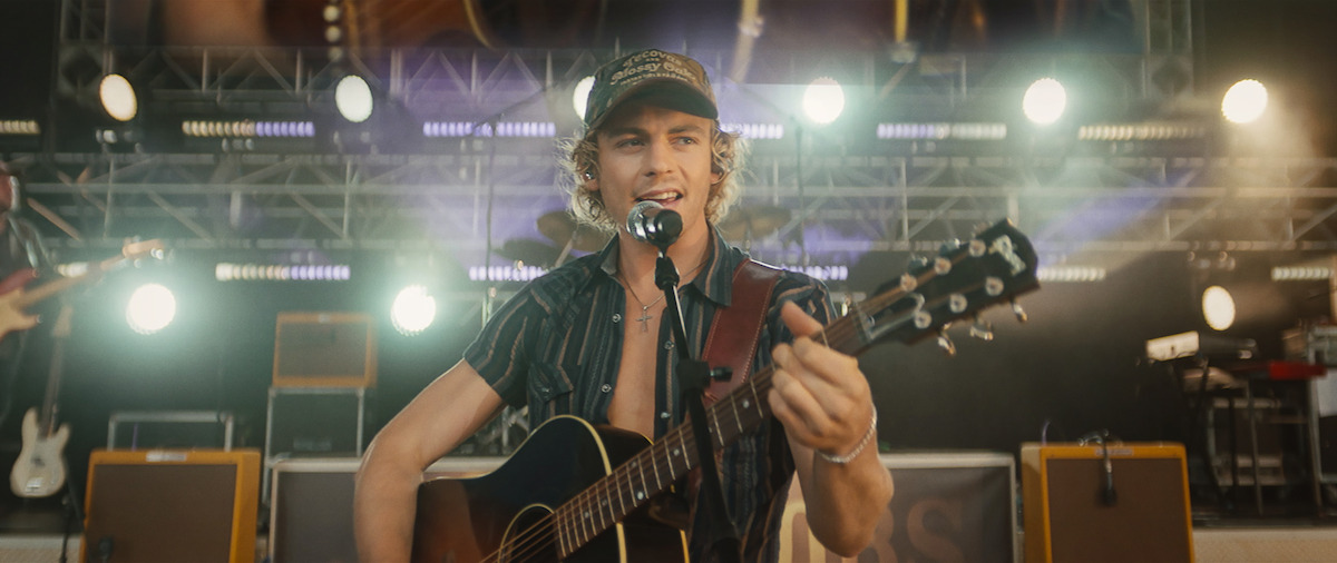Young male musician in a hat performs singing and playing guitar onstage at a concert, with stage lights, amplifiers, and instruments in the background.