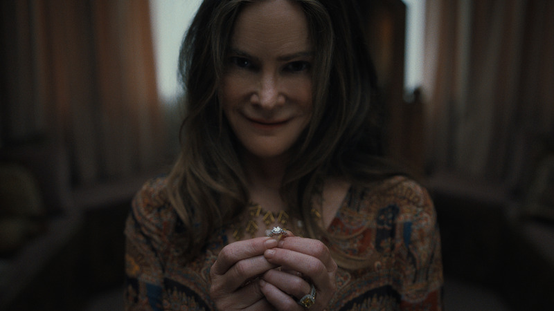 A woman with long brown hair and patterned blouse smiles subtly while holding a ring in both hands, in a dimly lit room with soft curtains and neutral furniture in the background.