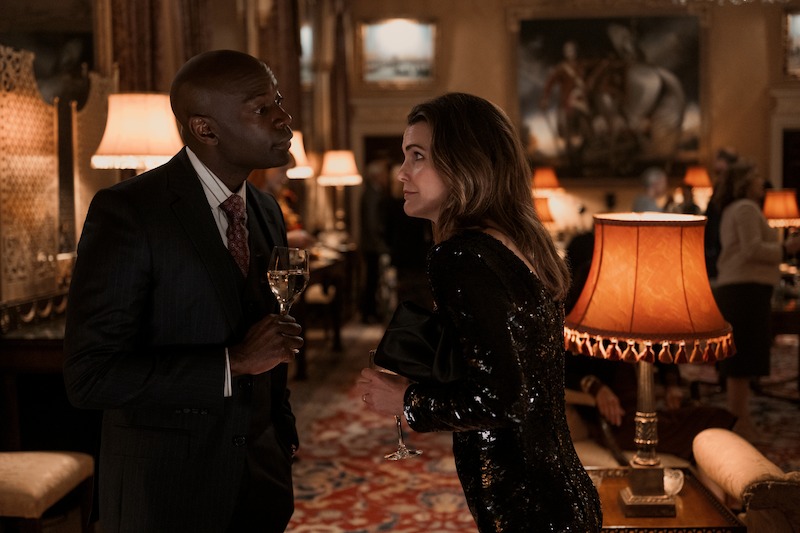 A man in a suit and a woman in a black sequin dress are holding drinks and having a conversation in an elegantly decorated, warmly lit room with lamps and paintings.