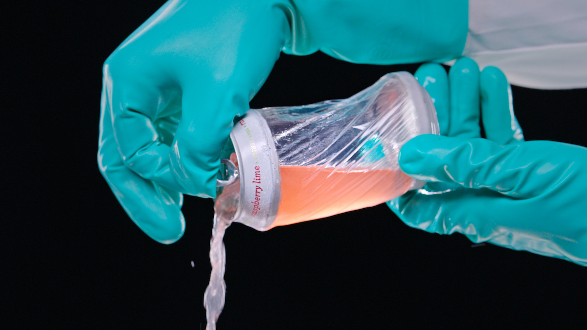 Hands in teal gloves pour liquid from a plastic-wrapped cup labeled “raspberry lime” against a dark background, likely in a controlled or experimental environment.