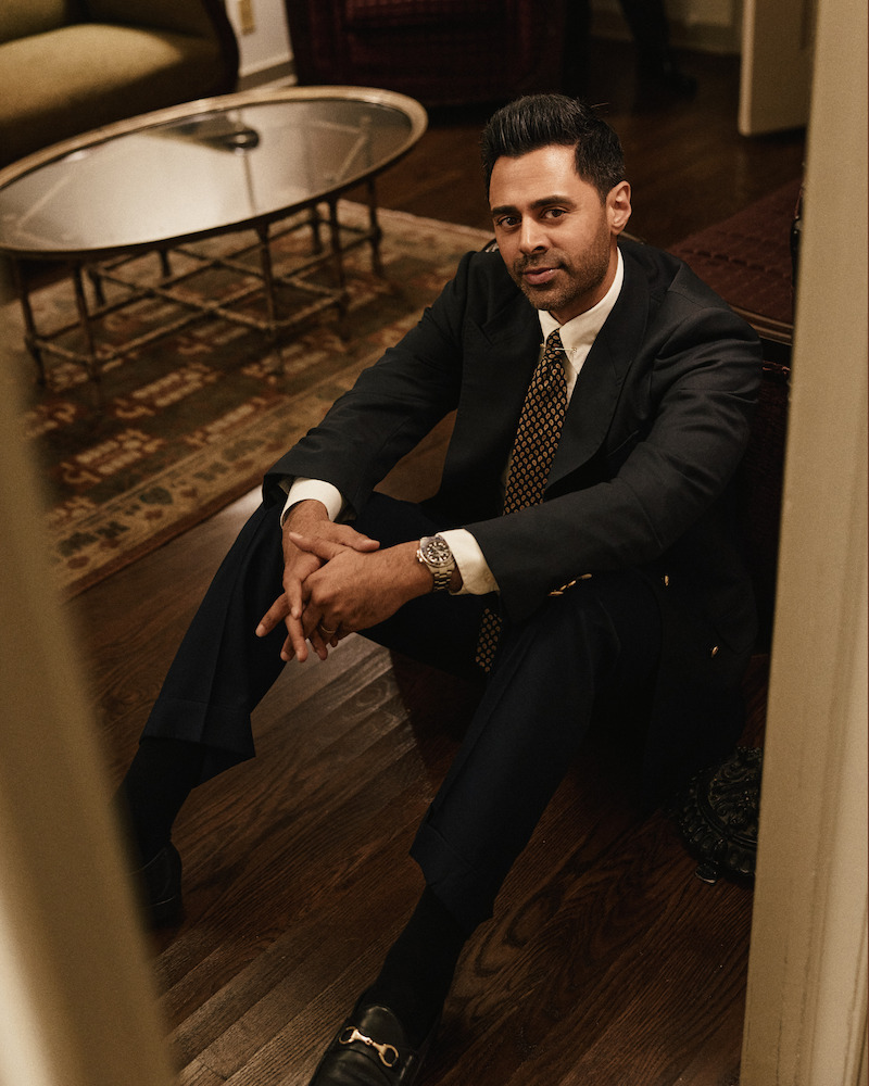 Man in a suit sits on a wooden floor in a warmly lit living room, near a round glass coffee table, patterned rug, and upholstered furniture, looking relaxed and thoughtful.