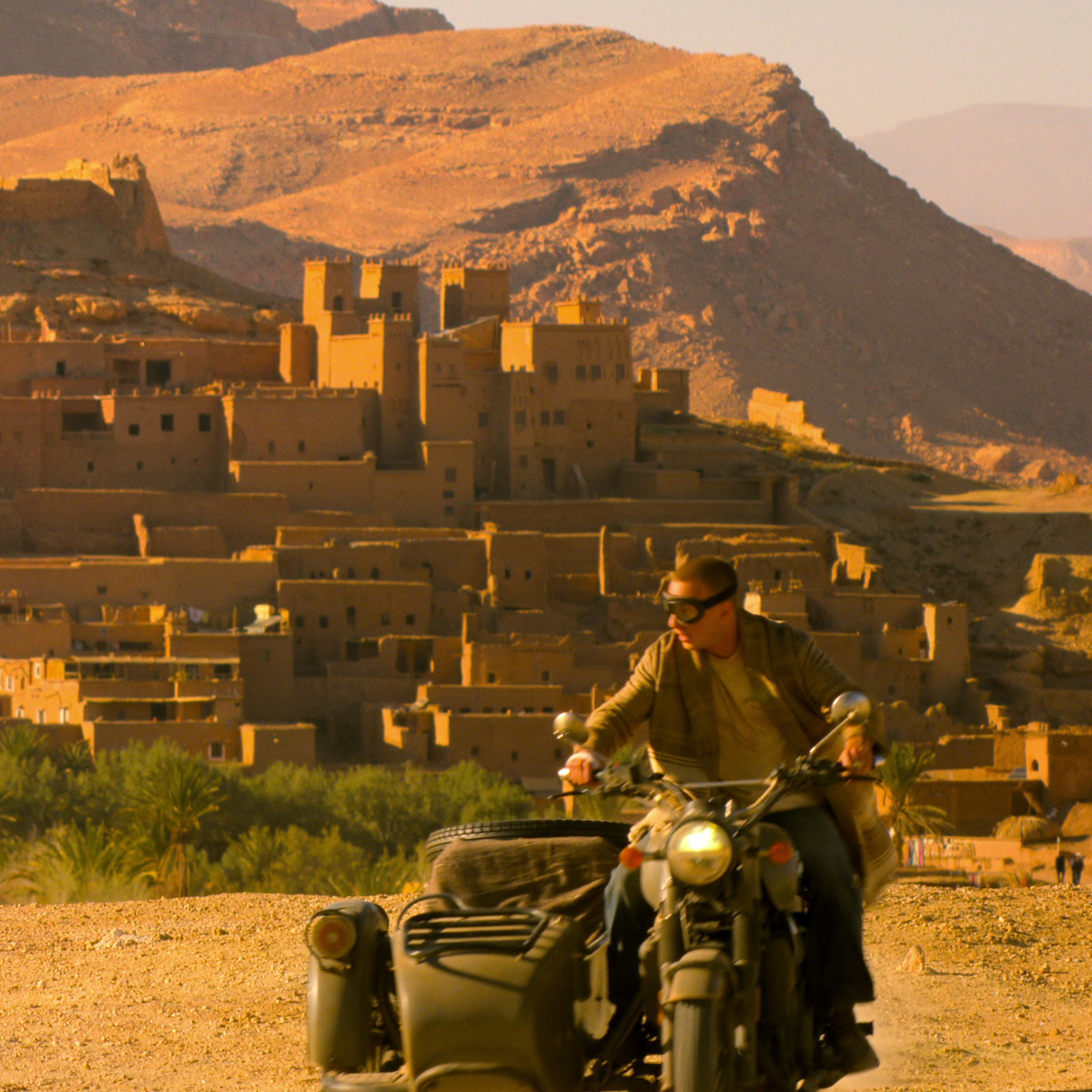 Homem pilotando uma motocicleta com sidecar em uma estrada empoeirada em frente a um vilarejo histórico no deserto, cercado de montanhas áridas e banhado pela luz do sol.