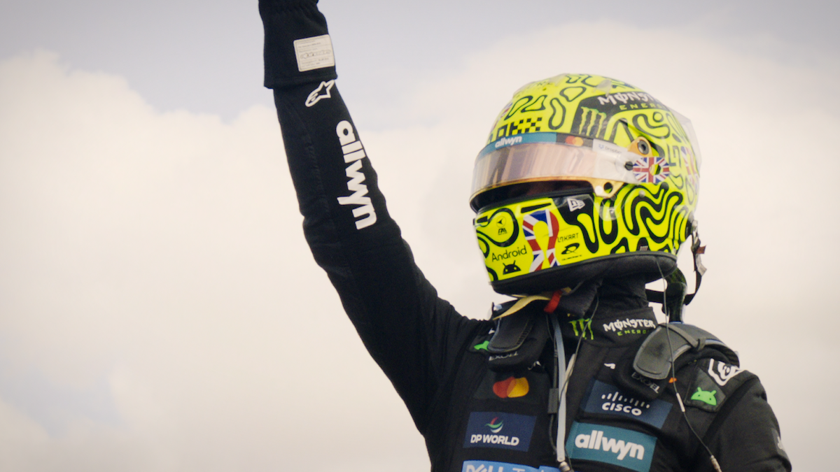 Race car driver in a colorful helmet and black suit raises arm in celebration against a cloudy sky, with sponsor logos visible on the driver's clothing and gear, capturing a victory or triumphant moment outdoors.
