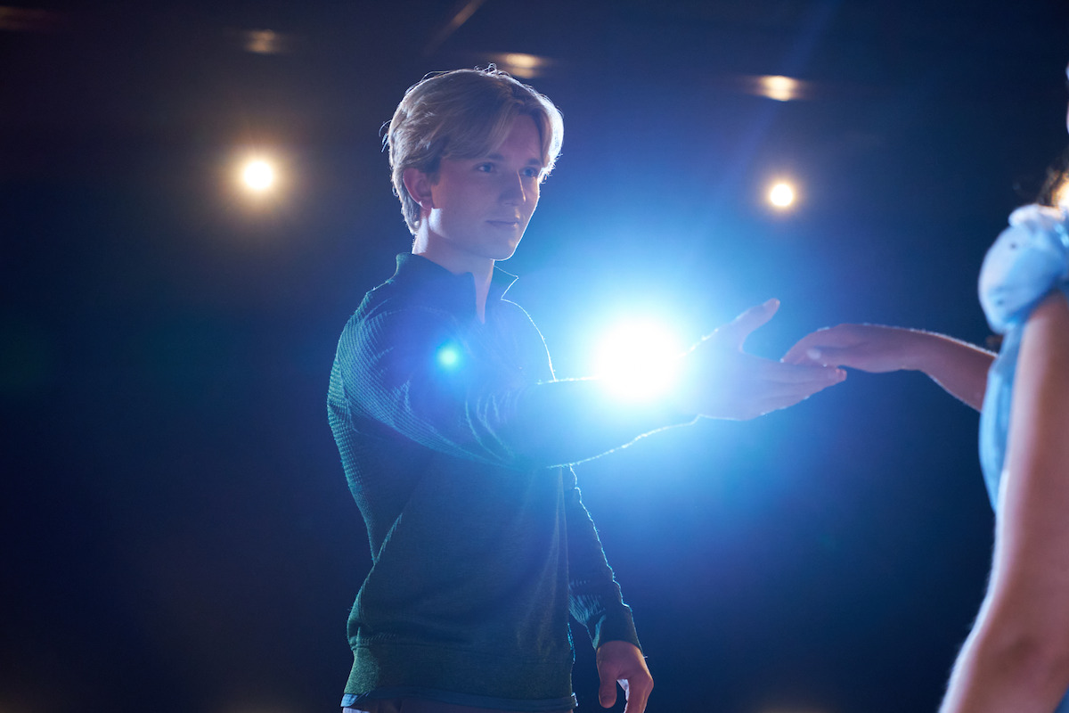A man reaching out his hand to a woman on a dimly lit stage, bright spotlight shining behind them, suggesting they are performing
