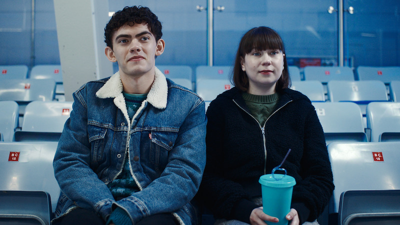 Two young people sitting in stadium seats, one holding a drink with a straw. They wear casual jackets and look forward, surrounded by empty seats in an indoor venue with blue tones.