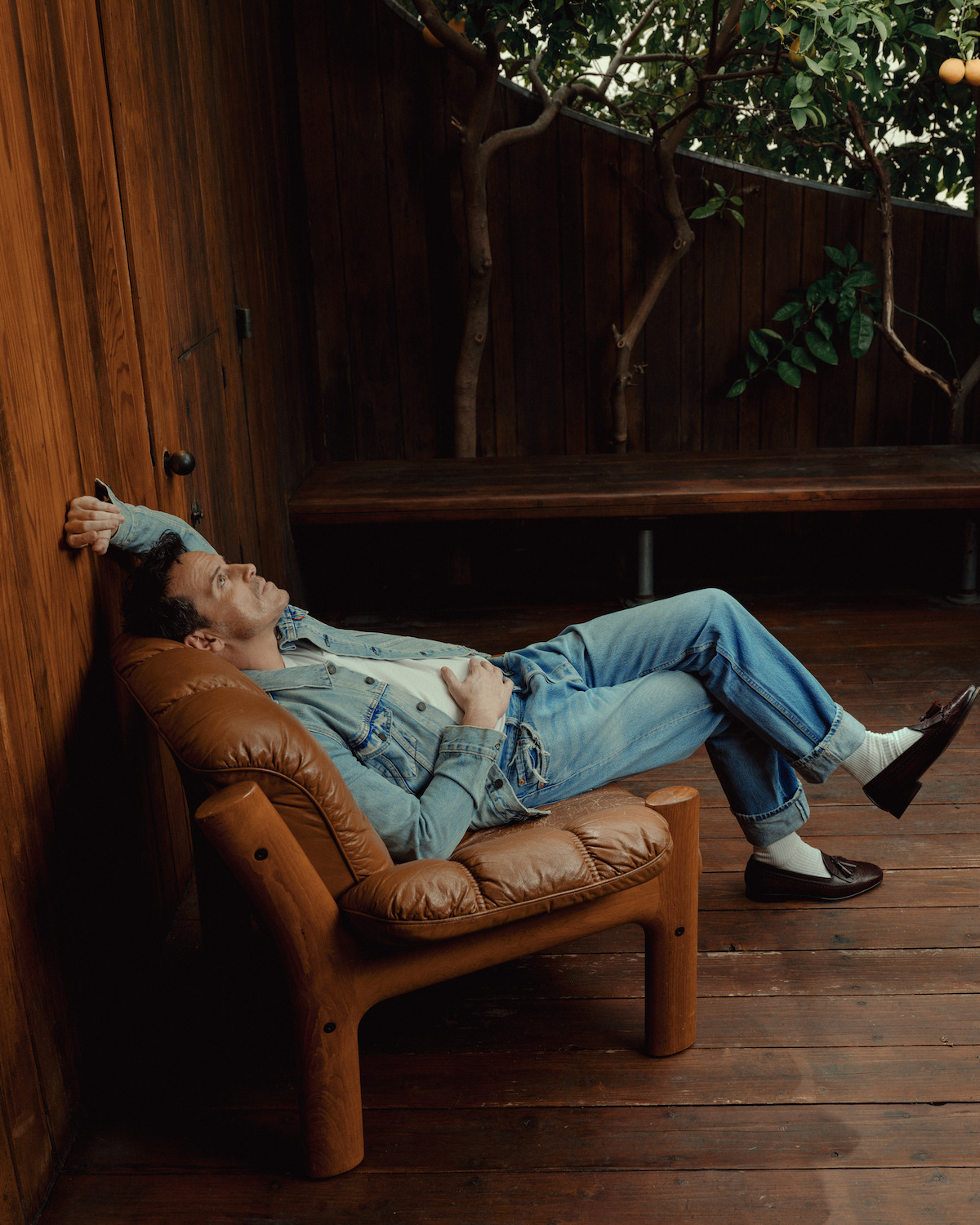 Andrew Scot wears a denim ensemble and looks pensively into some orange trees.