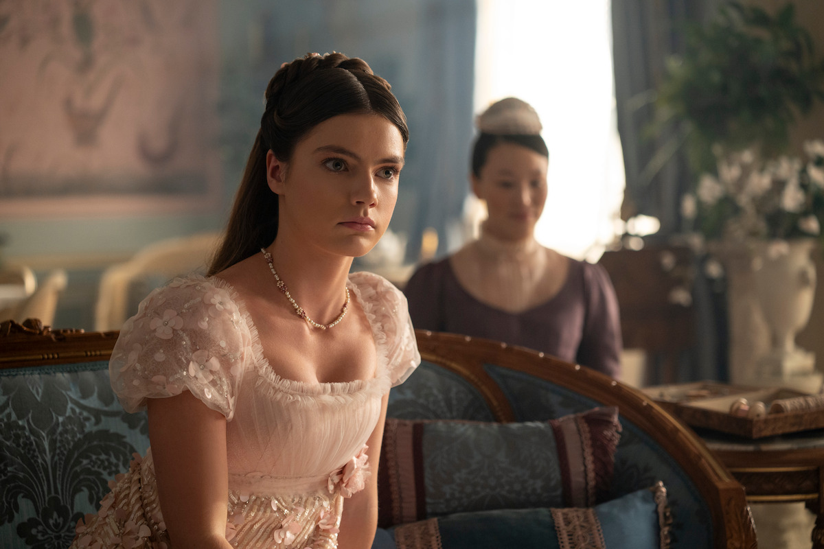 A young woman in an elegant, pale pink period dress sits on a blue patterned sofa in a richly decorated, softly lit room with another woman in the background. The setting suggests a historical or aristocratic environment.