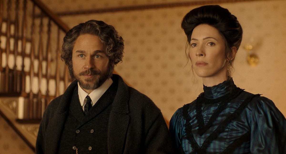 A man with curly hair and a beard stands beside a woman with dark hair in an old-fashioned, elegant dress, both in a warmly lit vintage interior with patterned wallpaper and a staircase in the background.