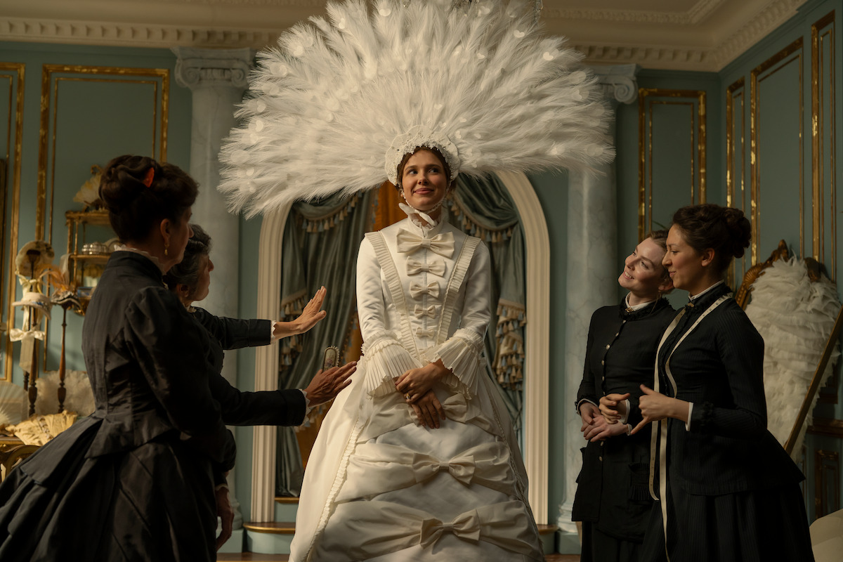 Woman in an elaborate white gown and large feathered headdress stands in an ornate, gold-trimmed room, surrounded by three women dressed in dark period clothing, as they adjust her outfit and admire her.
