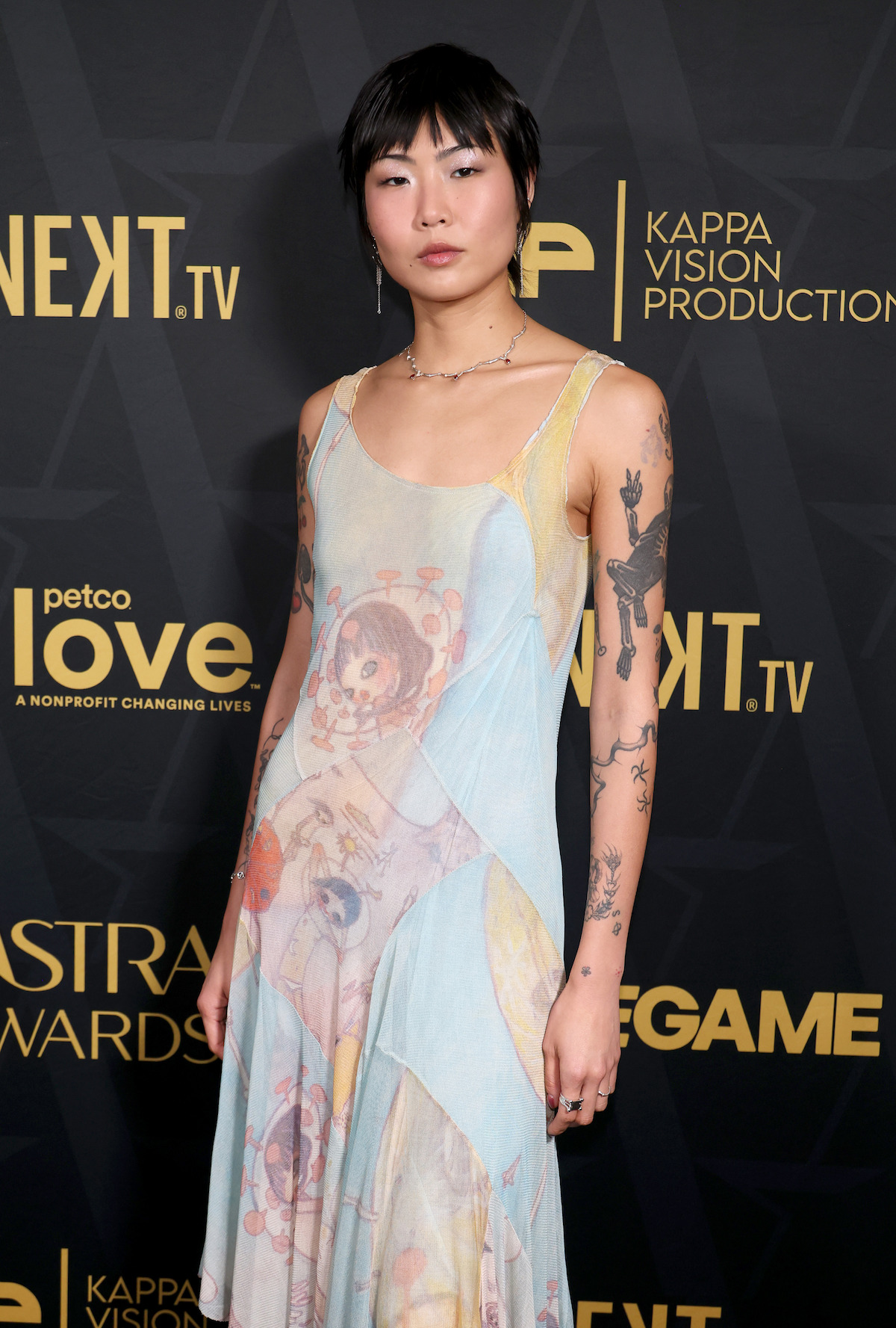 Person with short dark hair wearing a colorful, printed sleeveless dress, showing arm tattoos, standing in front of a dark backdrop with gold logos at an event or awards show.