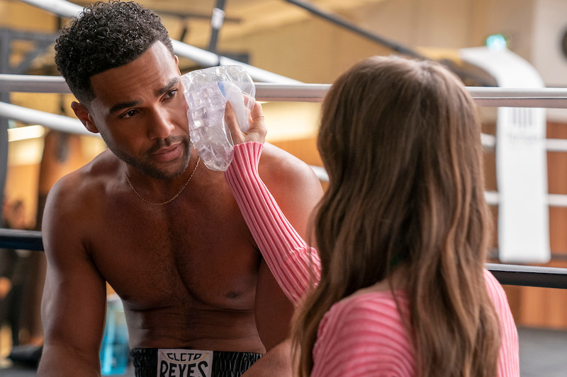 Lucien Laviscount as Alfie sits in a boxing ring while Lily Collins as Emily holds an ice pack to his face in Season 4 of 'Emily in Paris'