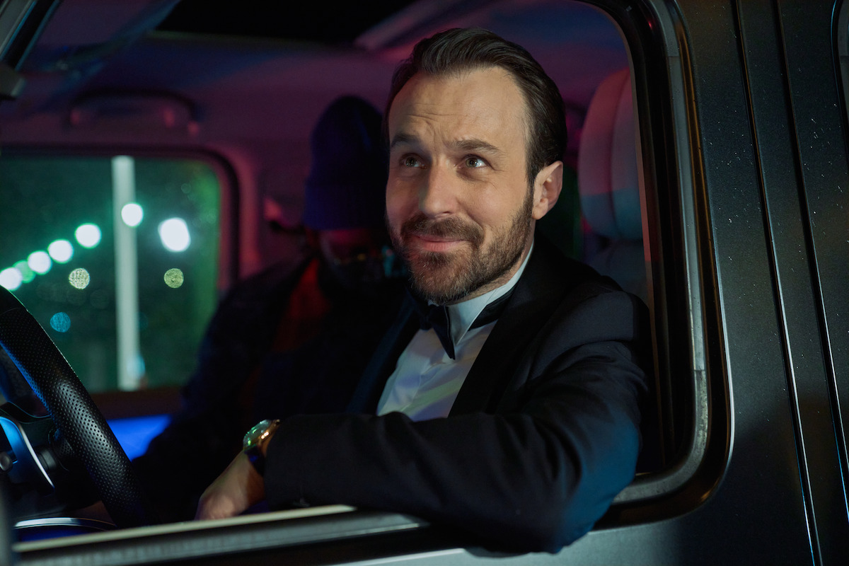 Man in a suit sitting in the driver's seat of a car at night, looking out the window and smiling, with city lights visible outside and a passenger in the background.