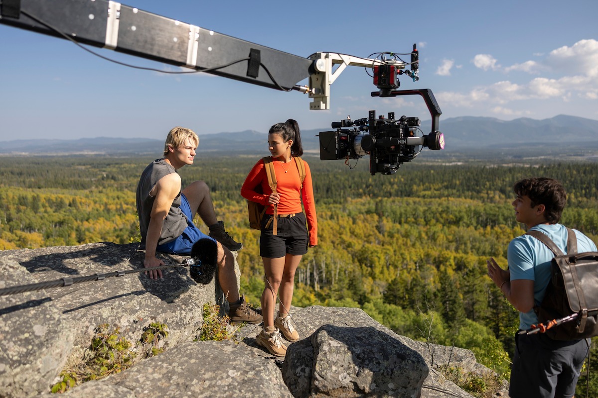 Three people with hiking gear on a rocky mountain ledge being filmed by a large camera crane, with a forest and distant mountains in the background under a blue sky.