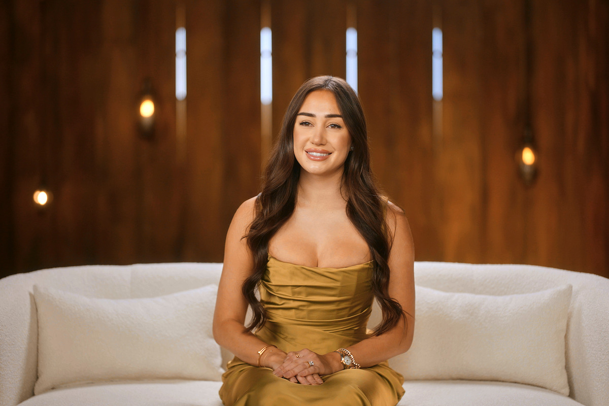 Woman with long brown hair in a gold dress sits on a white couch, smiling, against a warm wooden background with soft decorative lights and vertical slits, in a modern indoor setting.