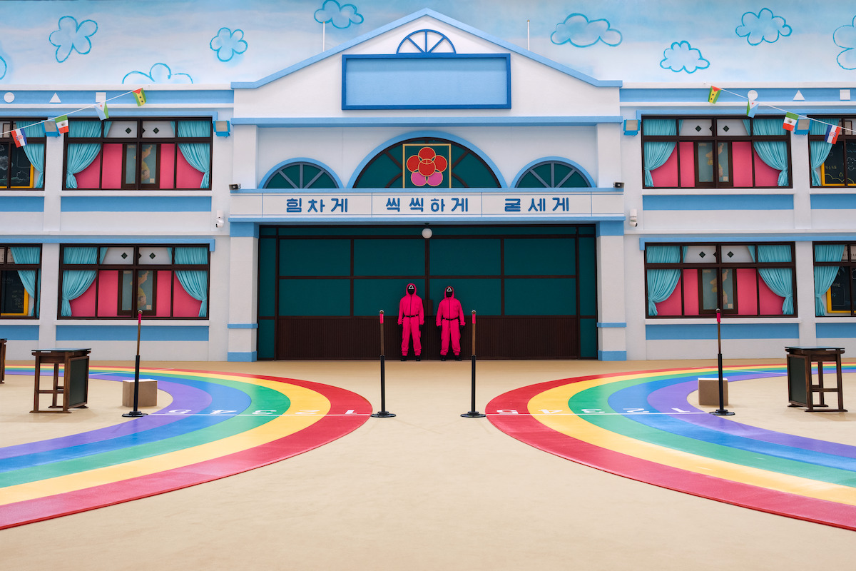 Two people in pink suits stand in front of a colorful, whimsical building with rainbow paths and playful decor, creating a surreal and playful yet controlled atmosphere.