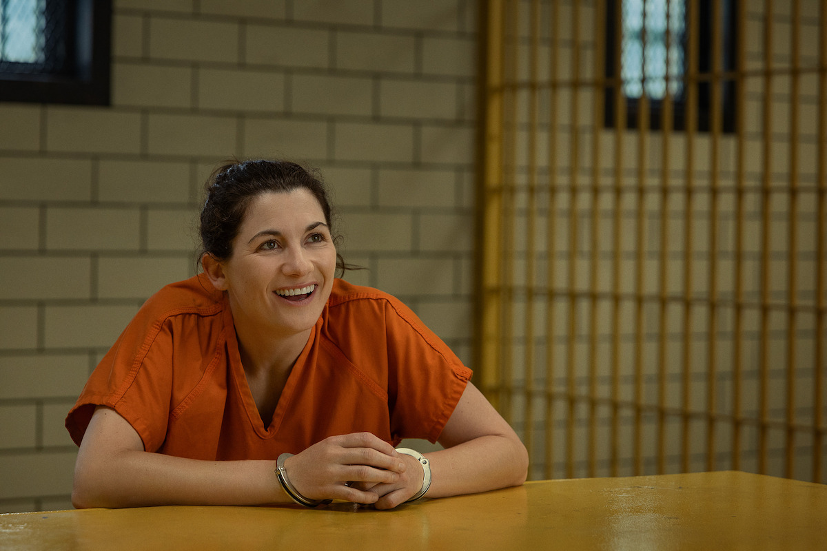 A woman in an orange prison uniform sits smiling at a table in a visitation area with tiled walls and barred windows, suggesting a jail or prison setting.
