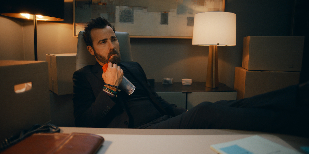 Man in a suit sitting with feet on desk, drinking from a bottle, surrounded by cardboard boxes in a dimly lit office with a table lamp, abstract art on wall, and paperwork on the desk.