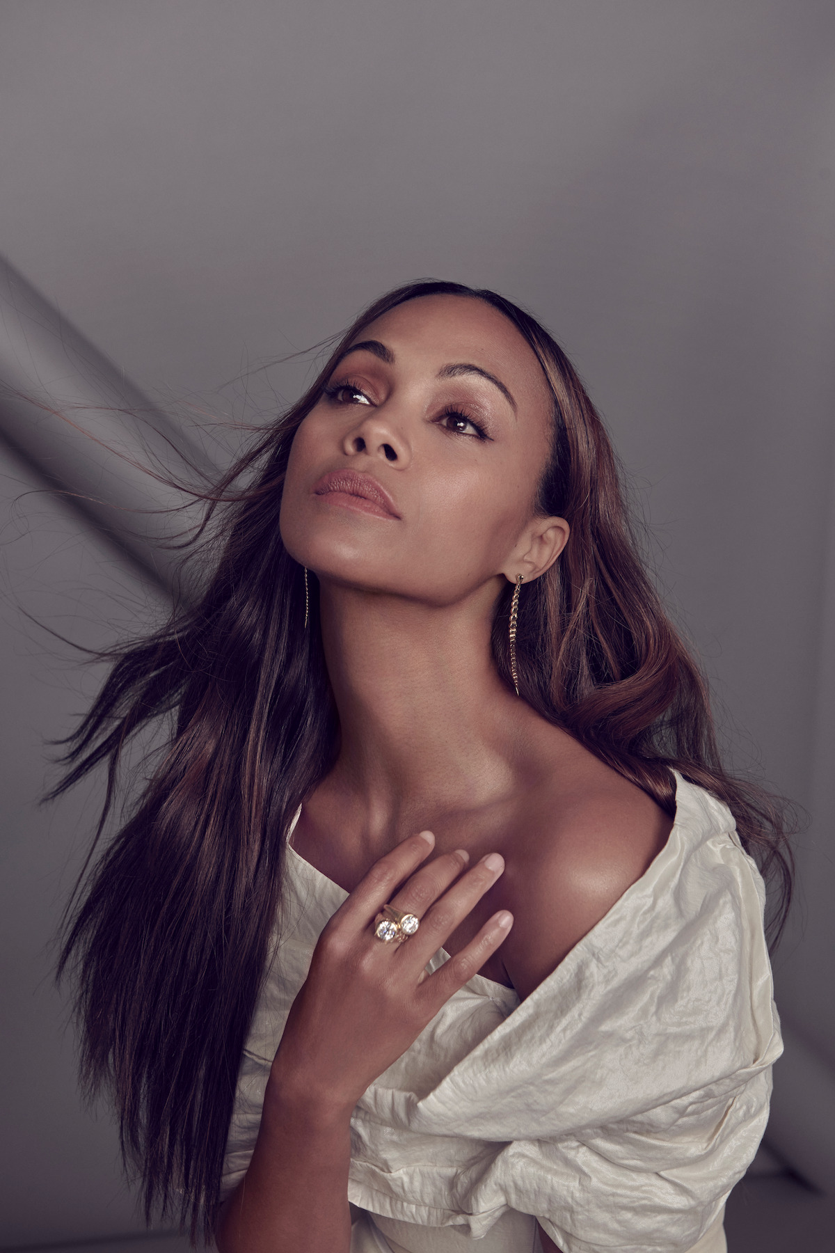 Zoe Saldaña looks up, away from the camera, with her hand on her collarbone and wearing a large diamond ring.