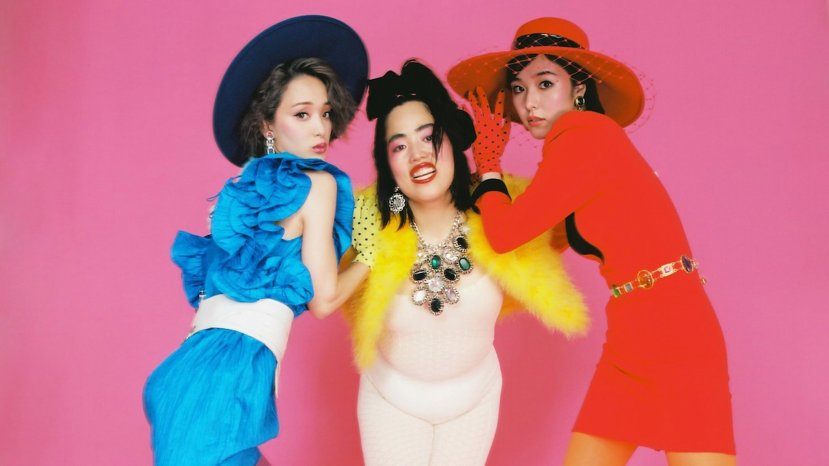 Yuriyan Retriever, Erika Karata, and Ayame Goriki pose wearing bright outfits and pose in front of a pink background