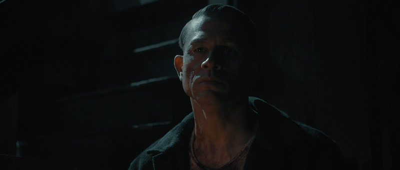 A man with a serious expression and visible scars on his face sits in a dimly lit, shadowy environment.