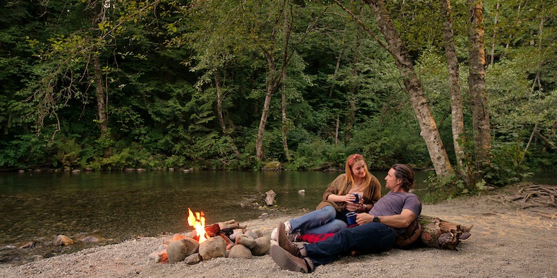 Alexandra Breckenridge as Mel Monroe and Martin Henderson as Jack Sheridan sit by a campfire near a lake  in Season 5 of ‘Virgin River.