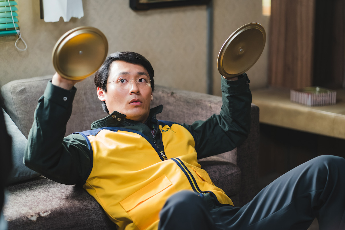 Surprised man in glasses and yellow vest sits on a couch holding two metal lids up, appearing startled, in a cozy indoor living room with warm lighting and casual decor.