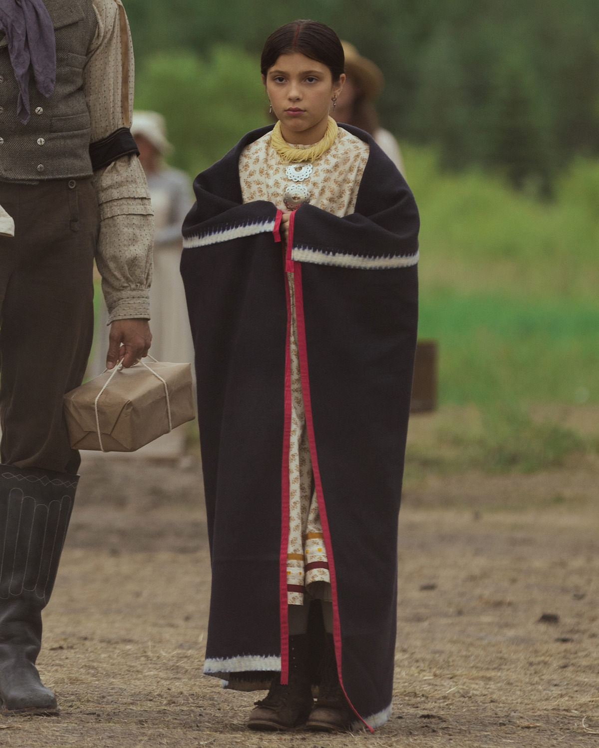 Young girl wrapped in a dark blanket with red trim, wearing a patterned dress and jewelry, stands outdoors on a dirt path next to an adult holding a package, with blurred greenery and people in the background.