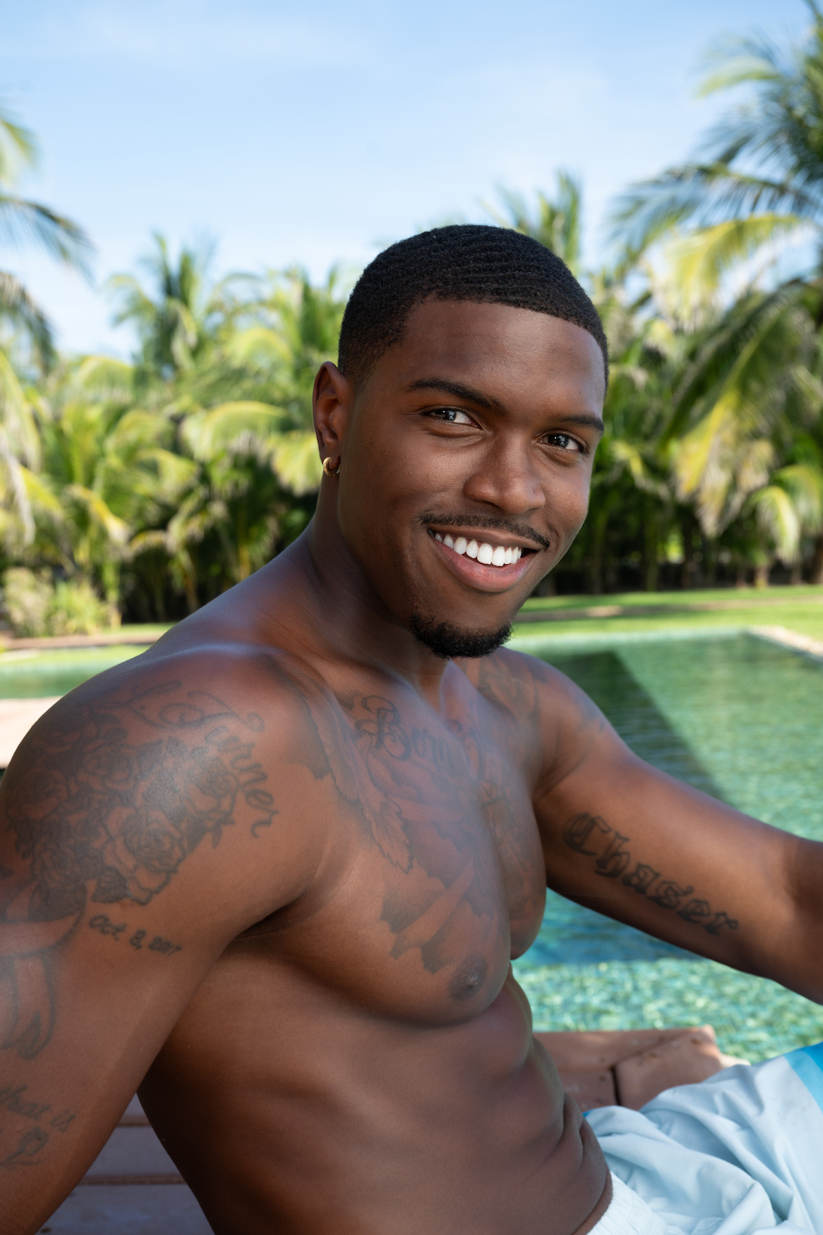 Smiling shirtless man with tattoos relaxing by a swimming pool surrounded by lush palm trees on a sunny day.