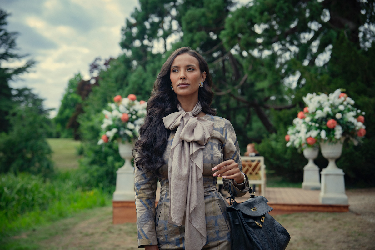 Woman with long dark hair in an elegant outfit and bow scarf stands outdoors in a lush garden with large floral arrangements and greenery, holding a black handbag on her arm.