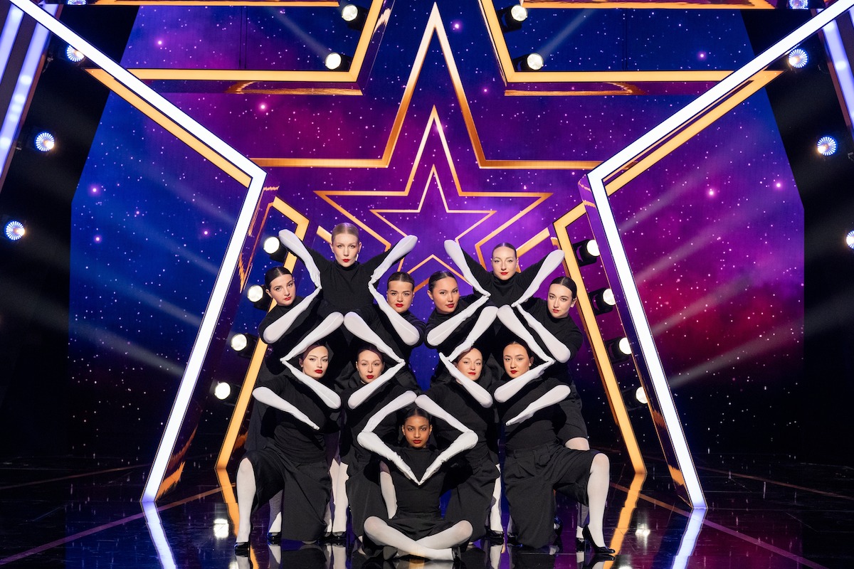 Ladymetry vestida con trajes blancos y negros realiza una formación sincronizada en un escenario con un gran fondo de estrellas iluminadas y vibrantes efectos de iluminación violeta y azul.