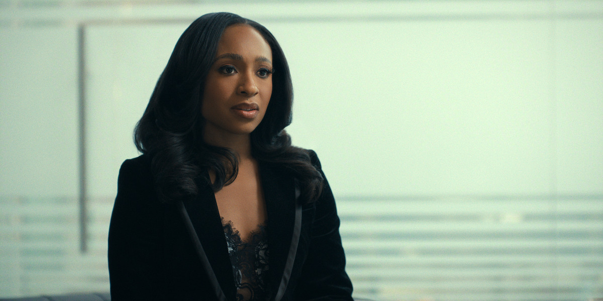 Woman with long dark hair in a black blazer sits indoors, with a blurred modern office background and horizontal lines visible through glass, appearing thoughtful or engaged in conversation.
