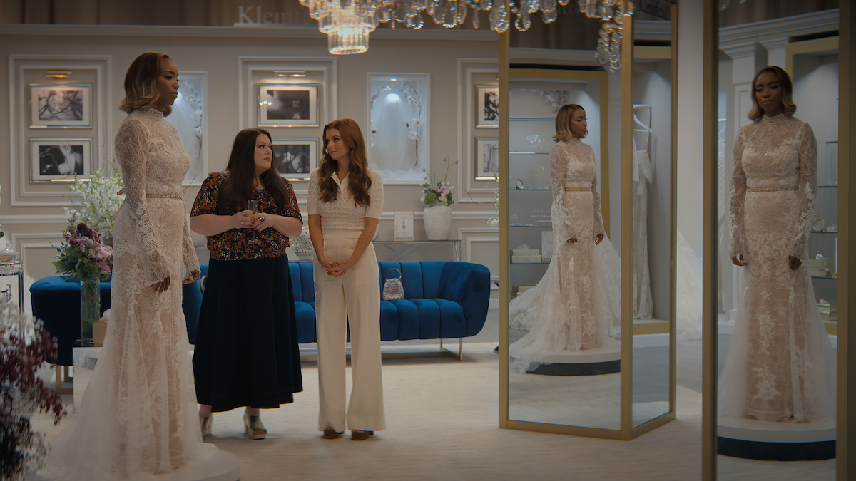 Three women in a stylish bridal shop, one wearing a wedding gown standing by mirrors, with elegant decor, framed photos on the wall, a blue couch, and chandeliers creating a classy atmosphere.