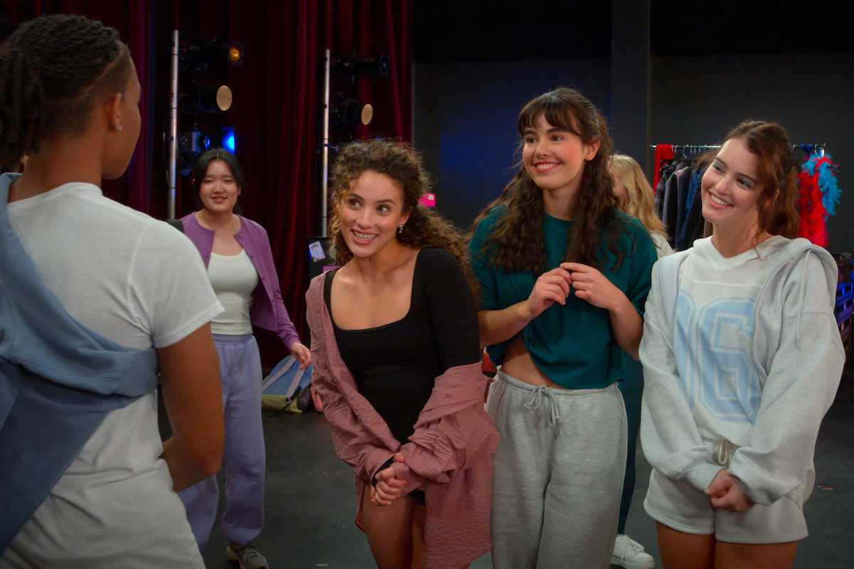 A group of young people in casual dance clothes stand together in a rehearsal studio with stage lights, red curtains, and costume racks in the background, socializing and smiling.