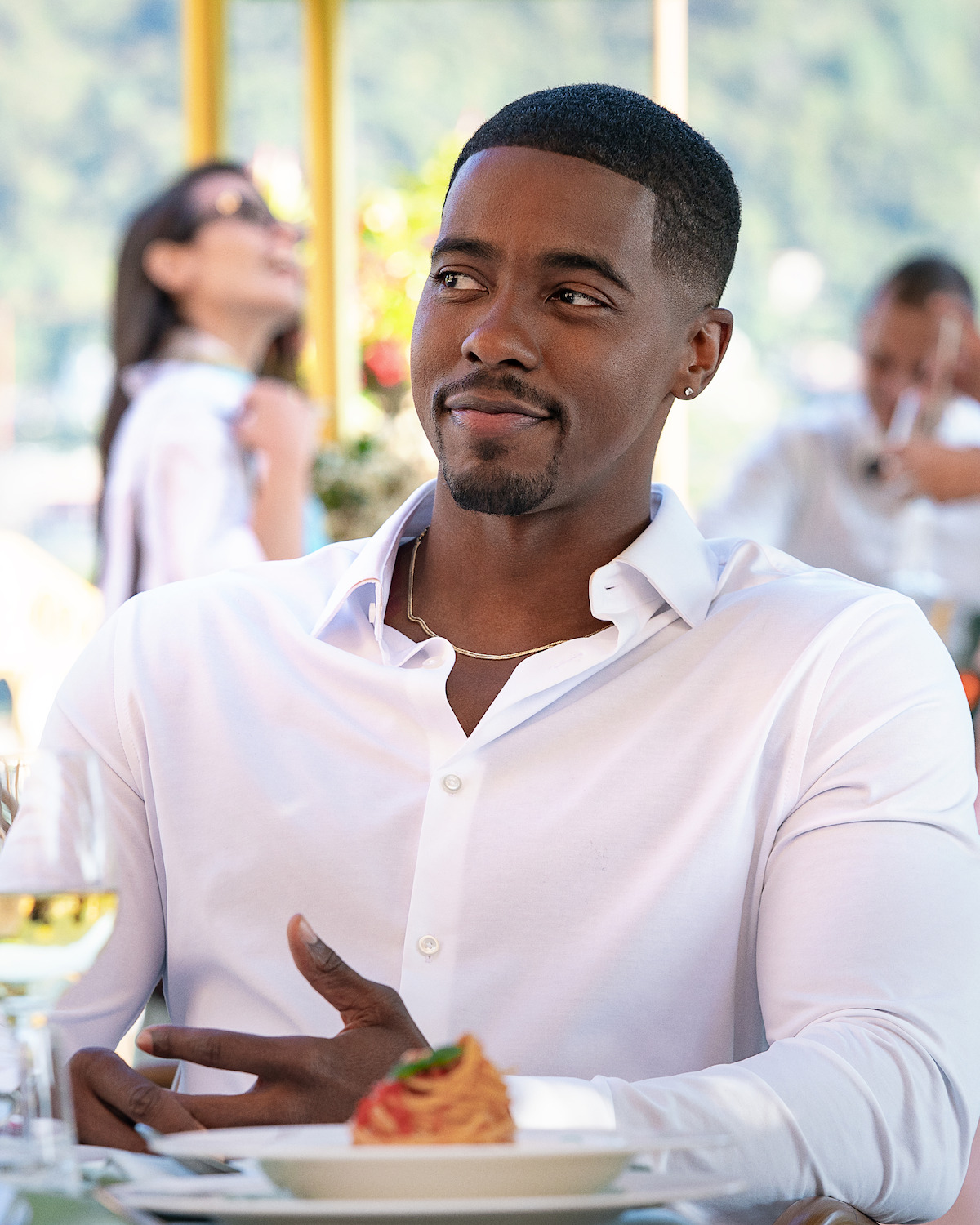 Man in a white shirt sitting at an outdoor restaurant table with food, with blurred people and greenery in the background, enjoying a casual meal.