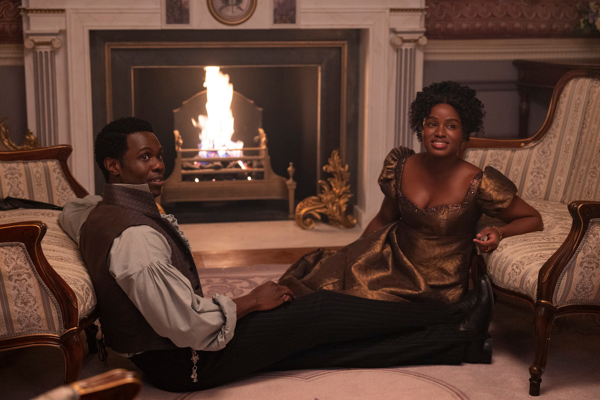 Two people in elegant period clothing sit on a luxurious living room floor by a fireplace, surrounded by ornate chairs and warm lighting, creating a cozy, historical atmosphere.