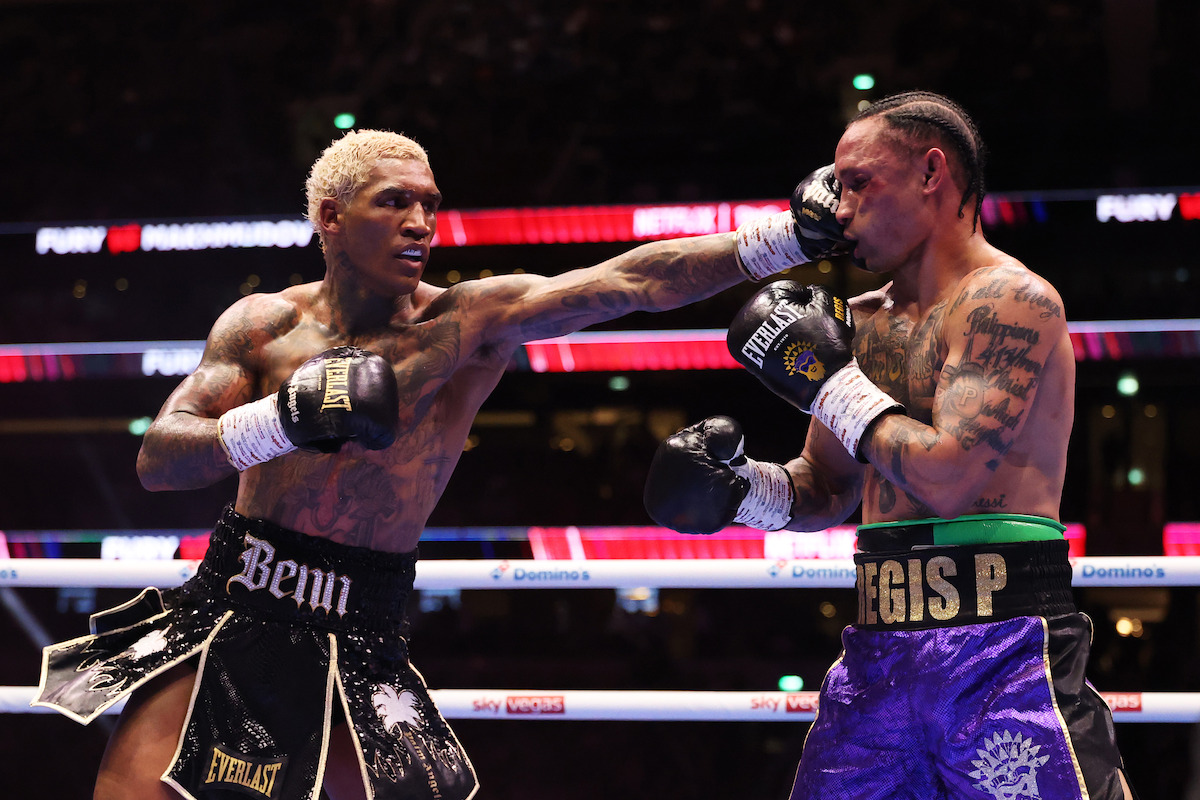 Two male boxers competing in a professional boxing ring, one landing a punch to the other's face. Both are wearing gloves, shorts, and are surrounded by bright arena lights and a cheering audience.