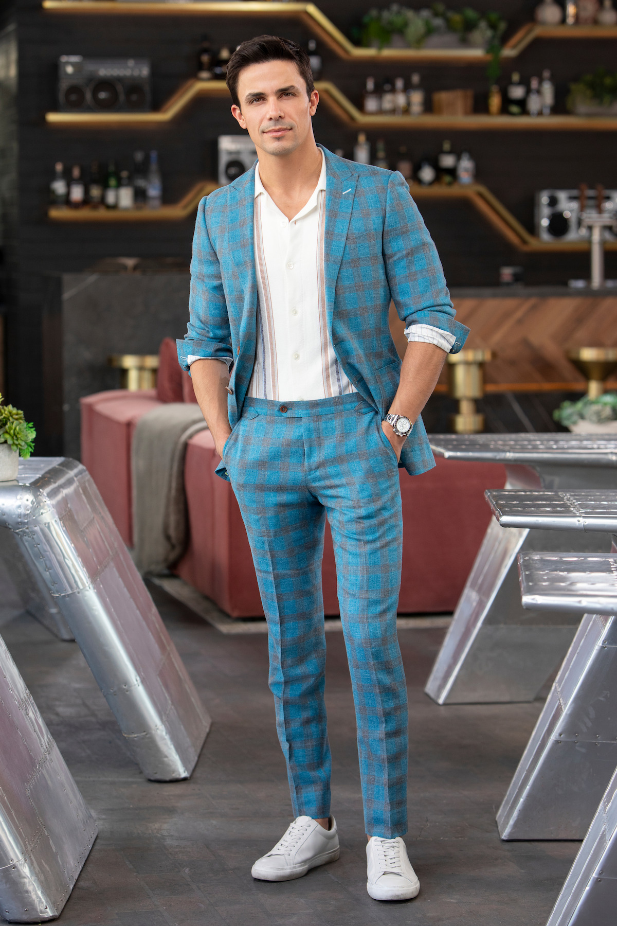A man in a blue plaid suit stands in a stylish, modern bar or lounge with geometric metallic tables, red couches, and shelves of bottles, exuding a confident and relaxed mood.