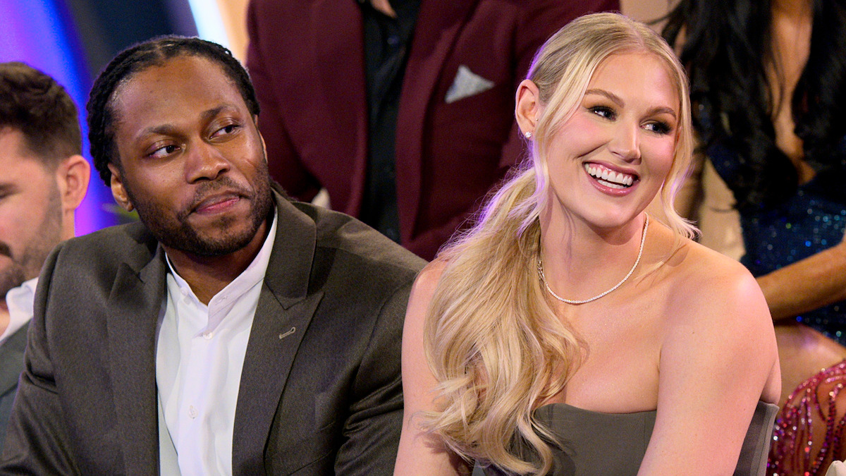 Man and woman dressed formally, sitting close together and smiling in a well-lit indoor setting with people seated around them, suggesting a social or television event.
