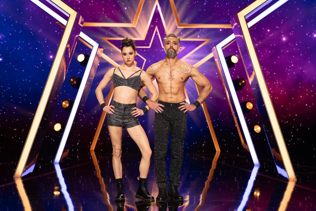 Duo Vespertilio in sparkly performance outfits stand confidently on a shiny stage with a star-shaped neon backdrop and purple starry lights, ready for a talent show performance.