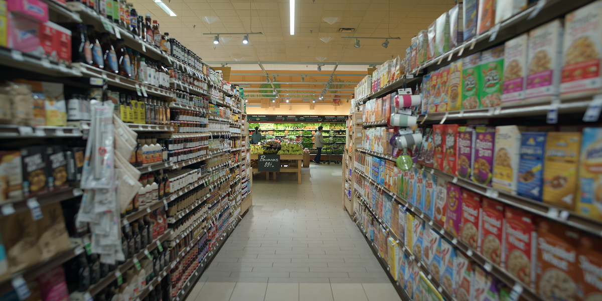 A grocery store aisle with shelves stocked with cereal on the right and sauces on the left, leading toward the produce section in a well-lit supermarket environment.