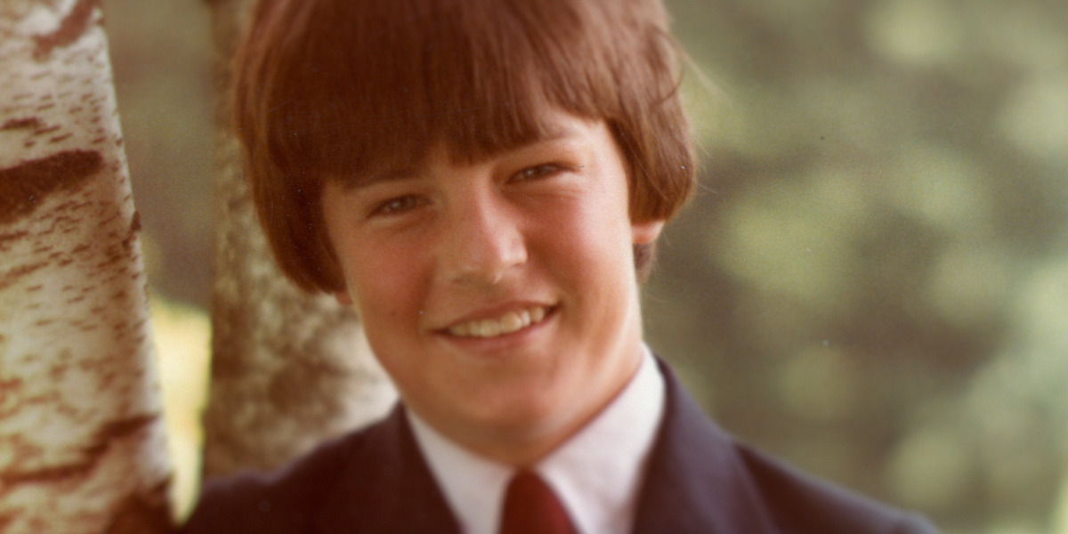 Smiling boy with brown hair in a suit and tie, standing outdoors next to a birch tree, blurred green natural background.