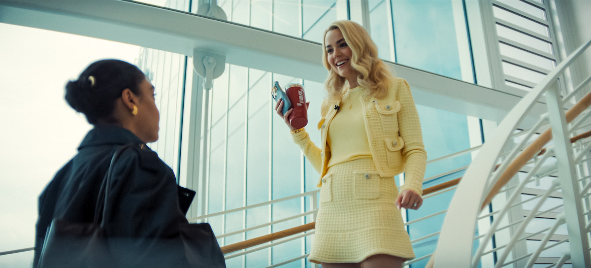 Two women talk in a modern glass building; one stands on stairs in a yellow outfit, smiling and holding coffee.