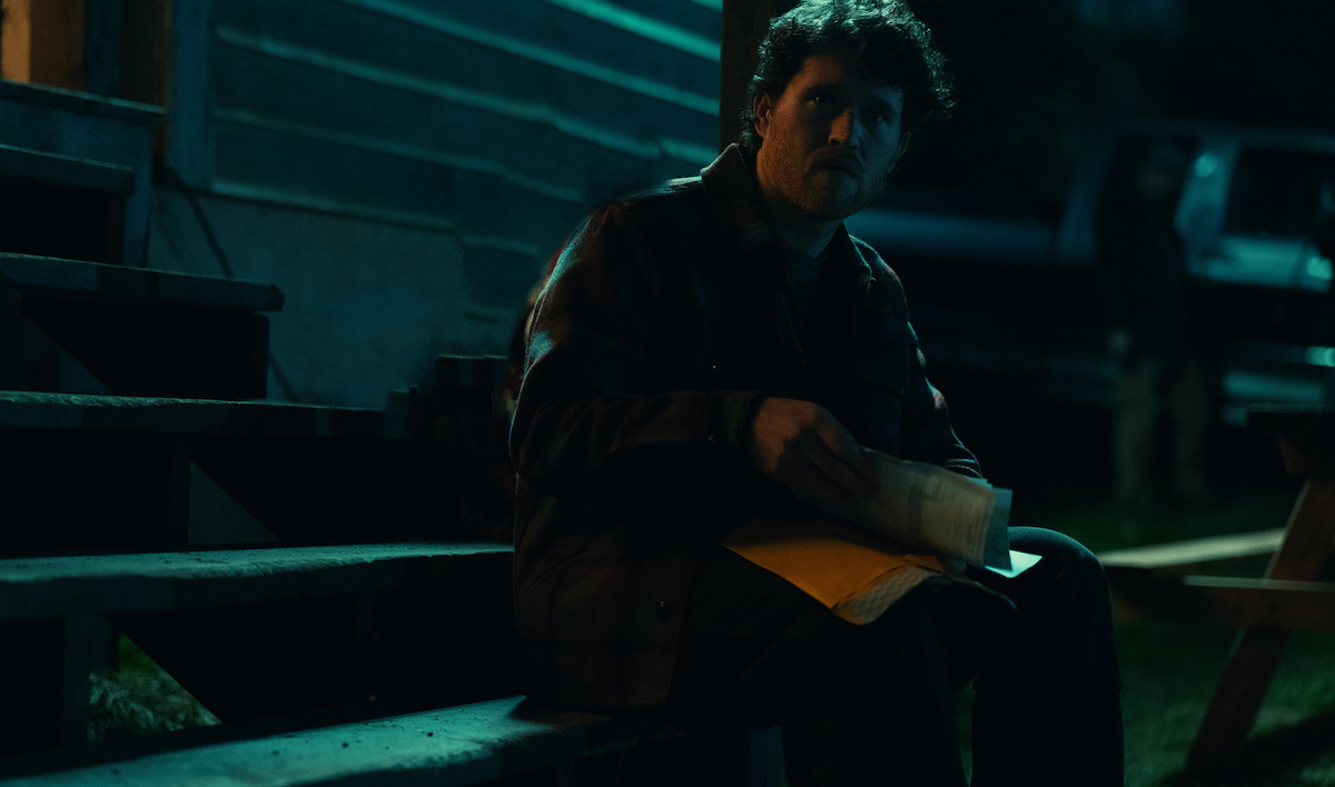 Um homem sentado em degraus de madeira em um ambiente externo à noite, segurando papéis e um envelope, parcialmente iluminado por luz azulada, ao lado de um edifício com um veículo estacionado e uma mesa de piquenique no plano de fundo com iluminação baixa.