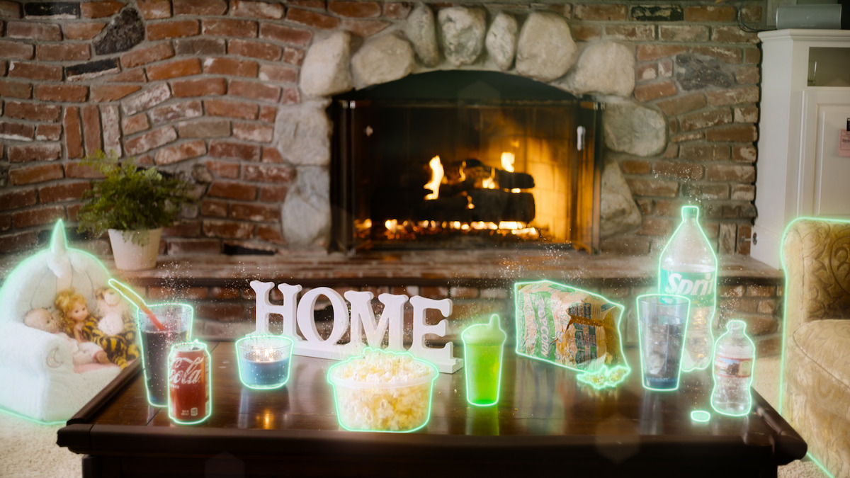 A cozy living room with a brick fireplace, burning fire, table with popcorn, drinks, snacks, and glowing neon outlines, plush toys on a chair, and HOME sign on the table. Warm, inviting setting for relaxing or movie night at home.