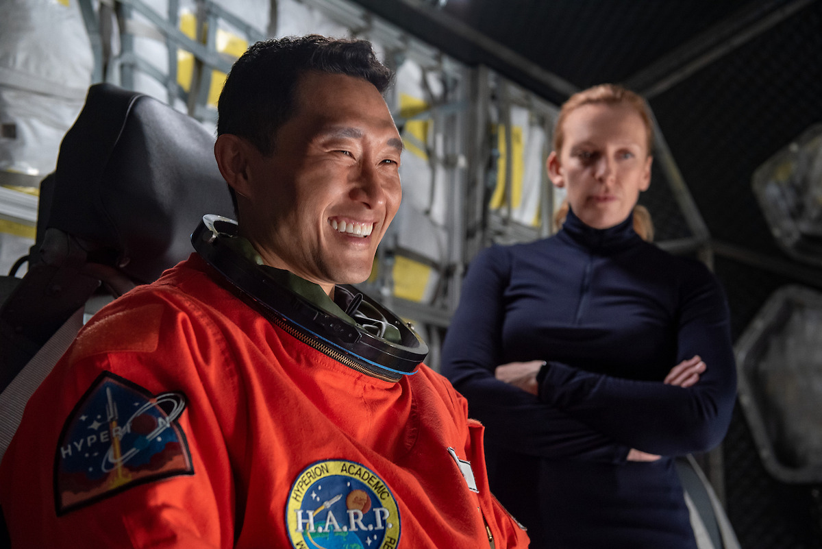 A man in an astronaut suit laughs as a woman watches him with arms folded