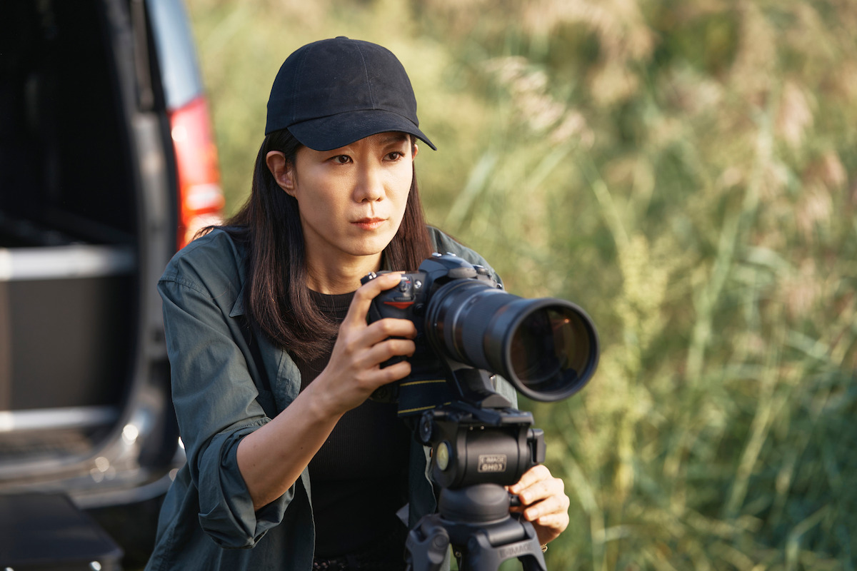 Wearing a black hat and a gray blouse, Jeon Hye-jin as Jang Hee-ju stands in front of a black SUV while taking a photo with a large camera on a tripod in a frame from the movie “Mission: Cross.”