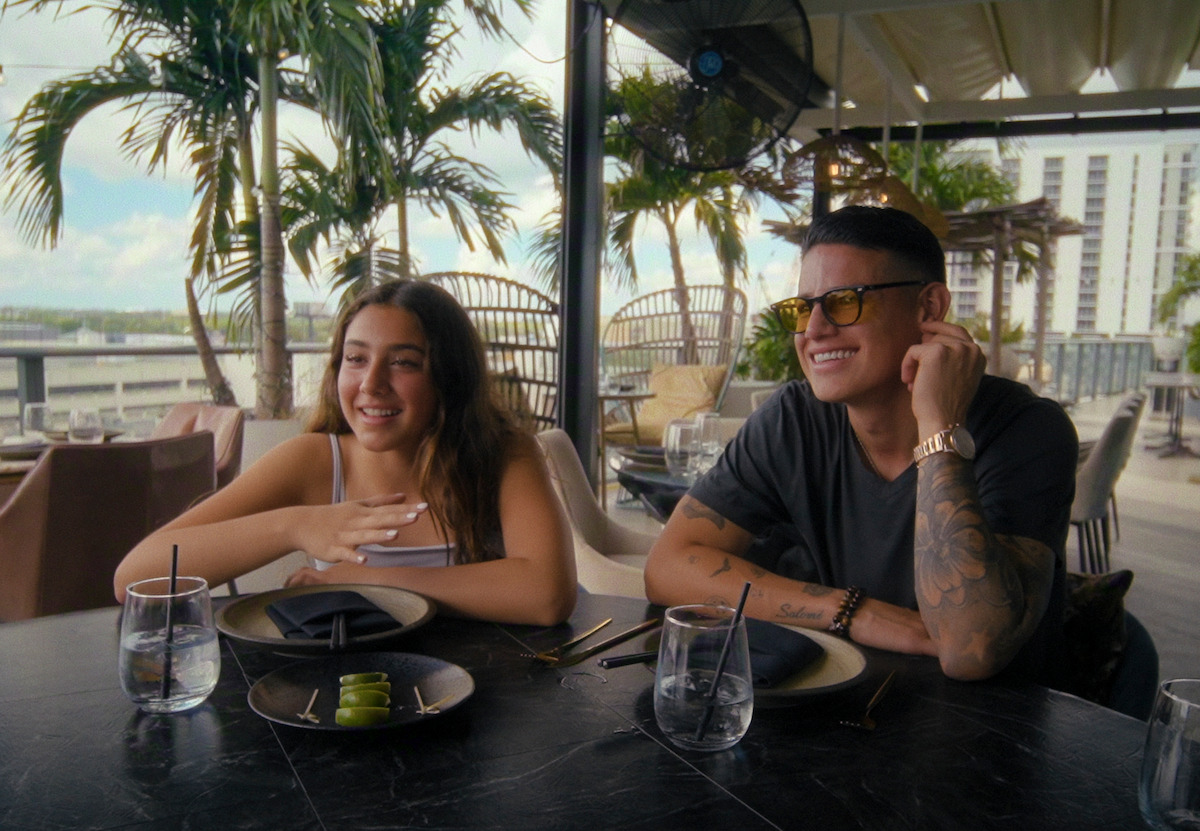 Two people sitting at an outdoor restaurant table, smiling and enjoying a meal, surrounded by palm trees and city buildings in the background under a partly cloudy sky.