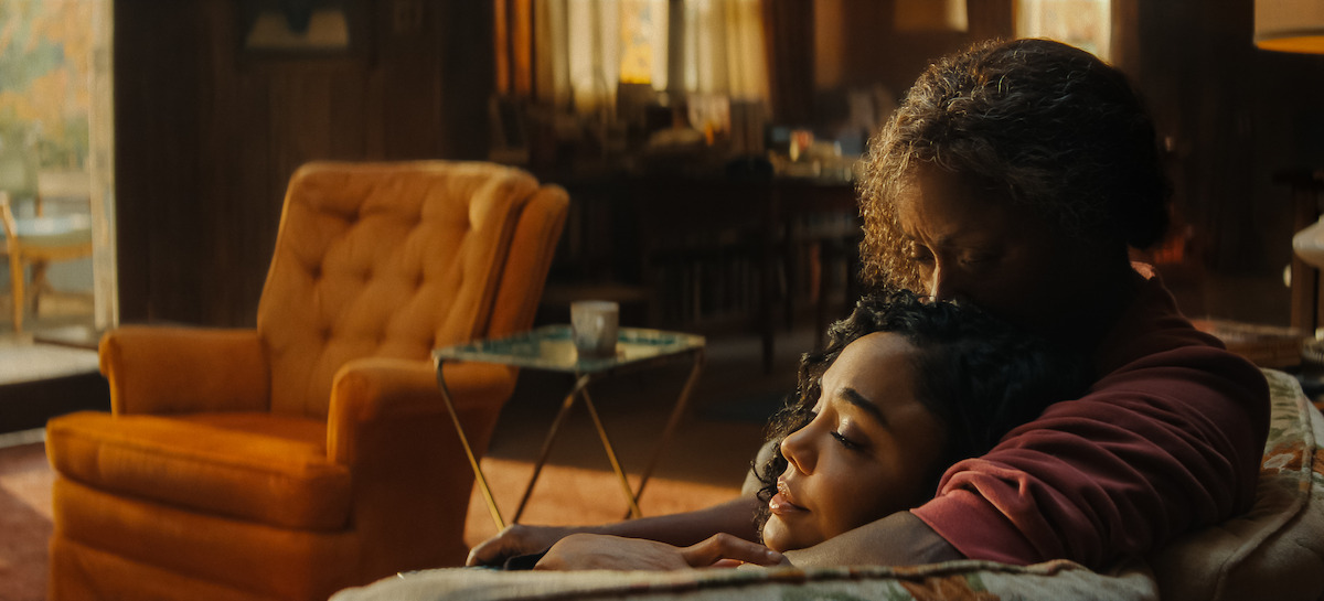 An older woman holds a young woman on a couch in a warmly lit, cozy living room with an orange armchair and soft natural light.
