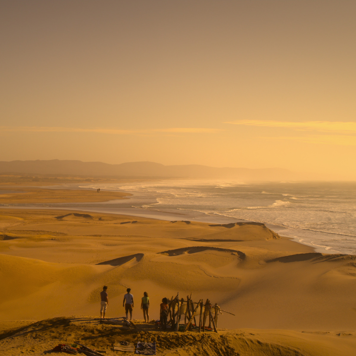 Grupo de pessoas em pé perto de uma estrutura rudimentar de madeira em dunas de areia com vista para uma praia extensa ao pôr do sol, com colinas distantes ao fundo sob um céu ensolarado.
