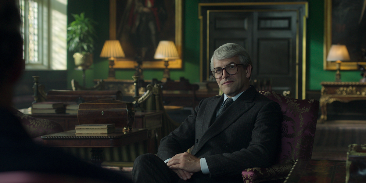 Jonny Lee Miller as John Major sitting in a chair